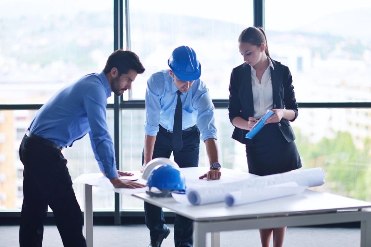 Three people in a room looking at plans on top of table.